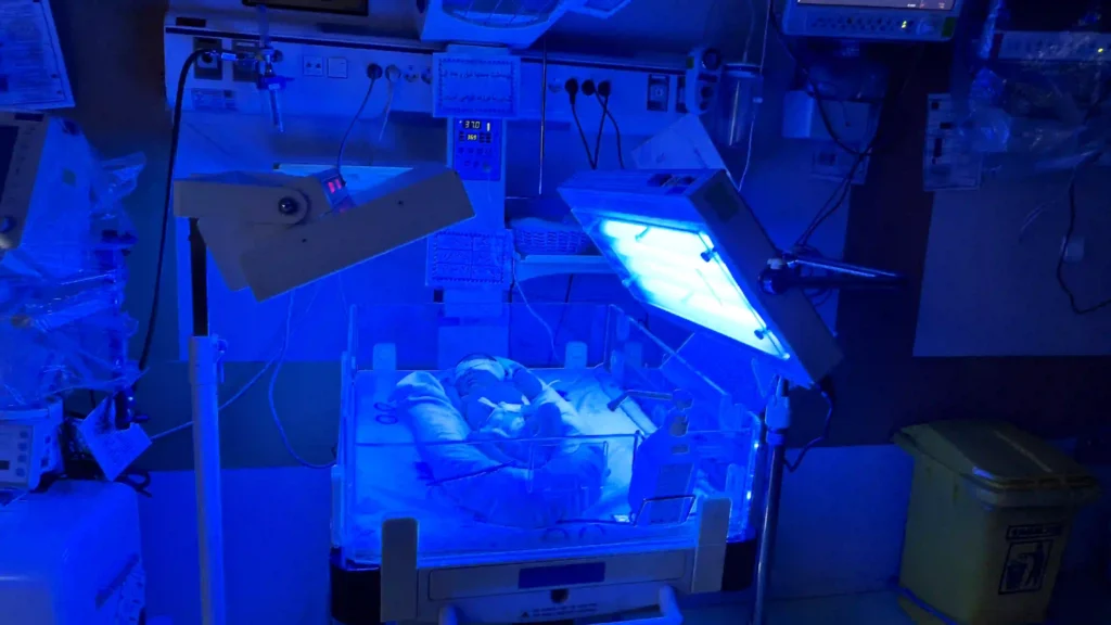 تصویری از بخش مراقبت‌های ویژه نوزادان (NICU) با نوزادان در تخت‌های گرم‌کننده و دستگاه‌های پزشکی متصل، تیم پرستاری در حال بررسی و مراقبت از نوزادان.